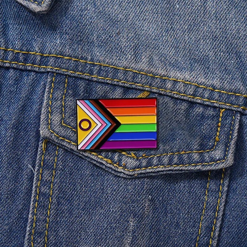 The Progress Pride Flag - Enamel Lapel Pin, featuring rainbow stripes and a chevron with black, brown, light blue, pink, and white accents, is attached to the pocket of a blue denim jacket as an inclusive pride accessory.