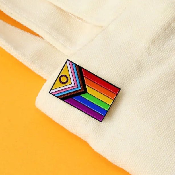 The Progress Pride Flag - Enamel Lapel Pin, rectangular and attached to a white fabric, features the inclusive Progress Pride flag design with a hint of yellow background—ideal for showing pride and support.