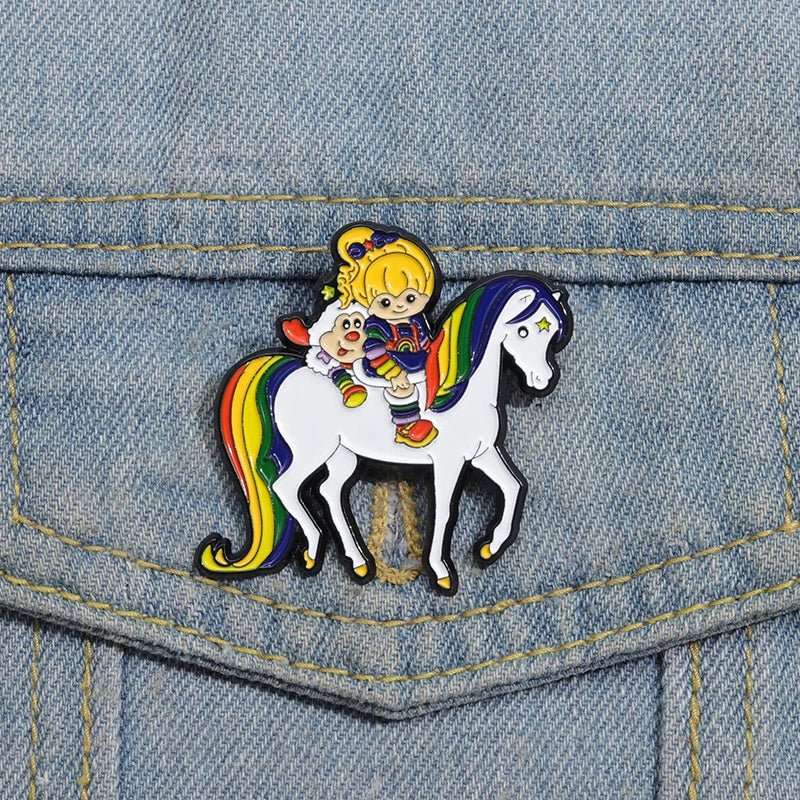 The Rainbow Brite Horse - Enamel Pin features a cartoon girl and creature riding a rainbow-maned white horse—ideal for Rainbow Brite fans or as a colorful pride accessory for your denim jacket or bag.