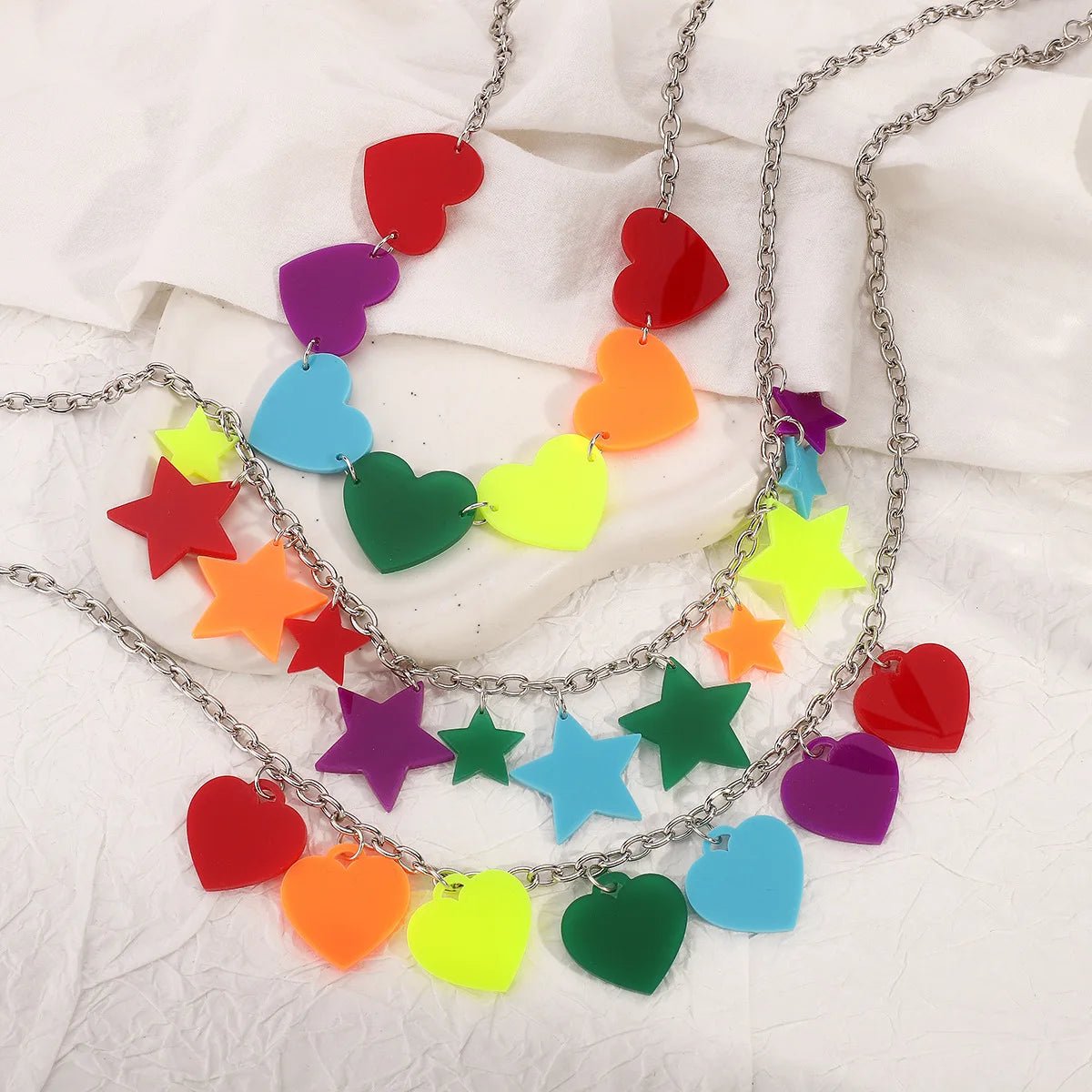 Three Rainbow Pride Acrylic Necklaces in 3 designs, featuring silver chains and colorful heart and star plastic charms in red, orange, yellow, green, blue, and purple, displayed on a white textured surface.