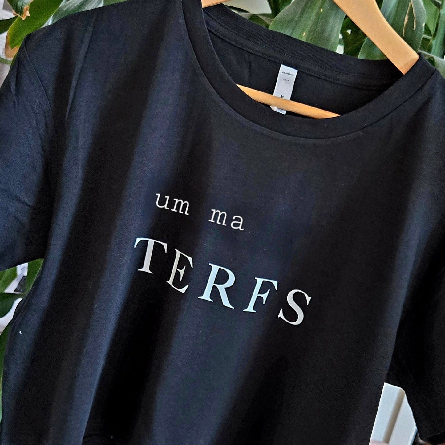 A black crop tee reading "um ma TERFS" in white text hangs on a hanger with green leaves in the background. Locally printed by a queer-owned small business.