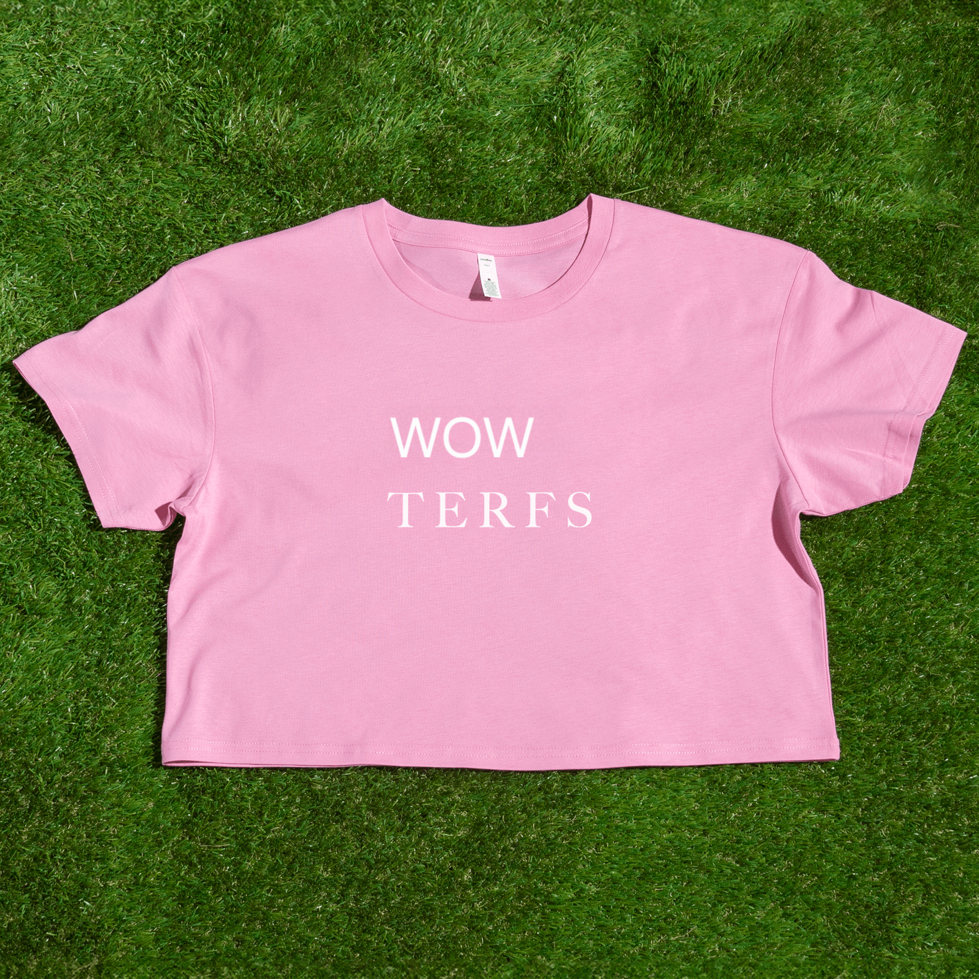 A locally printed pink TERFS crop tee with "WOW TERFS" in white, hand-printed by a queer-owned small business, shown on green grass.