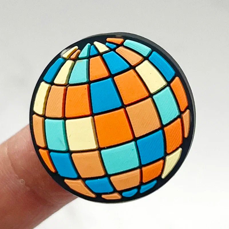 A finger holds a Retro Roller Skate theme Shoe Charm, featuring a spherical pin design with geometric patterns. The charm showcases alternating squares in orange, blue, yellow, and teal on a black background.