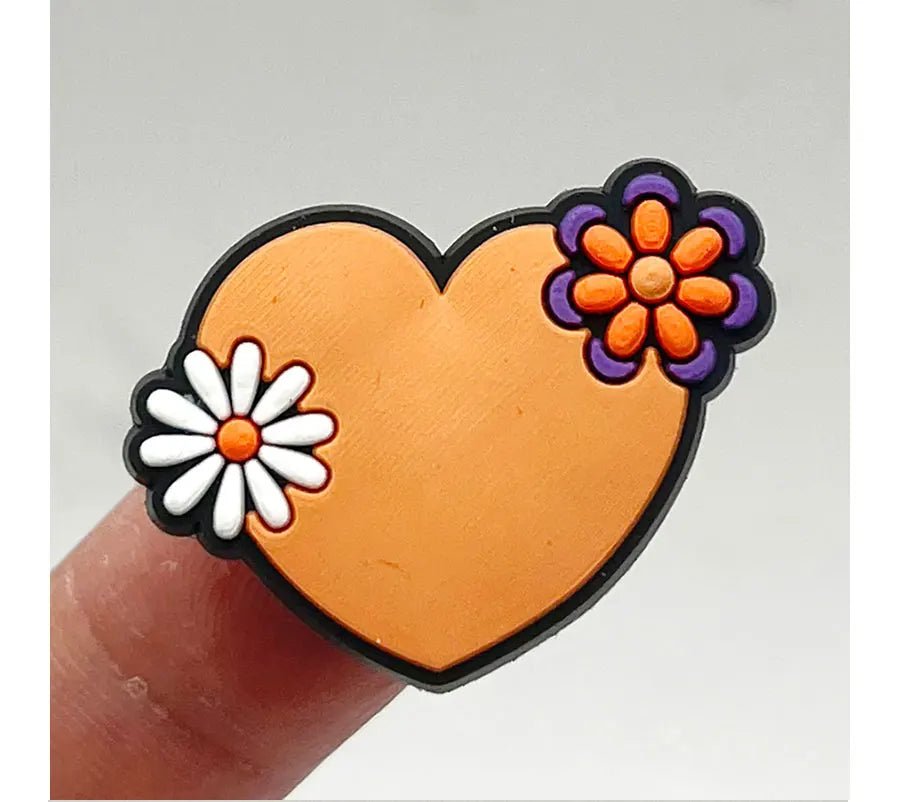 A Retro Roller Skate theme shoe charm, shaped like a heart and held between fingers, features a white daisy on the left and a purple and orange flower on the right. The plain gray backdrop highlights its vibrant colors.