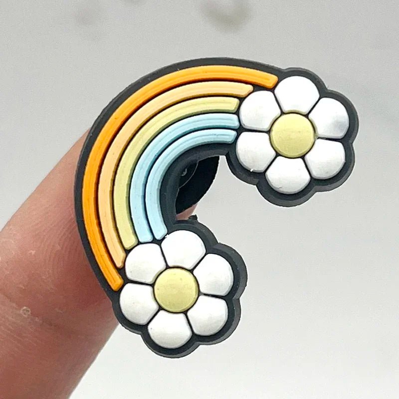 A finger holds a retro roller skate-themed rubber pin shaped like a rainbow with four stripes in orange, yellow, light blue, and white. This charming accessory features two white flowers with yellow centers at each end of its colorful arch.
