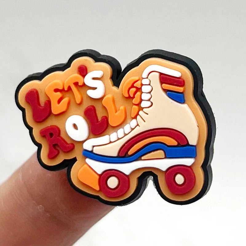 The Retro Roller Skate theme Shoe Charms feature the phrase "Let's Roll" in vibrant orange and red bubble letters alongside a vintage-style quad skate in shades of orange, cream, and red. Displayed between fingers against a plain backdrop, they capture a charming retro vibe.