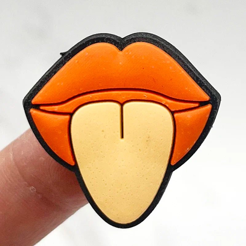 Close-up of a Retro Roller Skate theme Shoe Charm shaped like a stylized mouth with orange lips and a beige tongue sticking out, similar to an orange jibbitz charm. The pin rests on a person's finger against a white background.
