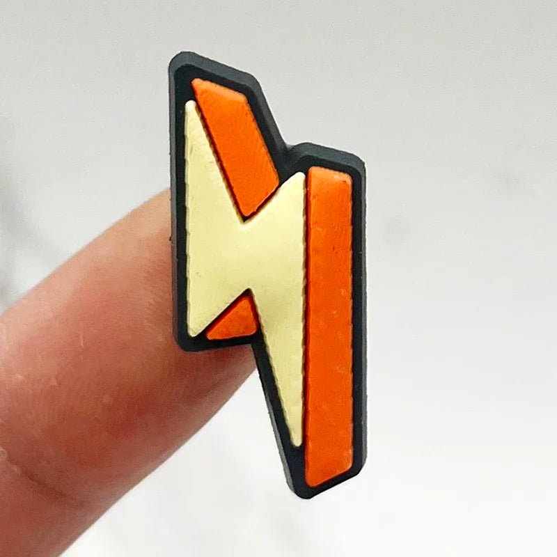 A Retro Roller Skate theme Shoe Charm shaped like a lightning bolt, featuring an orange and yellow design with a black outline, is held between two fingers against a light gray background.