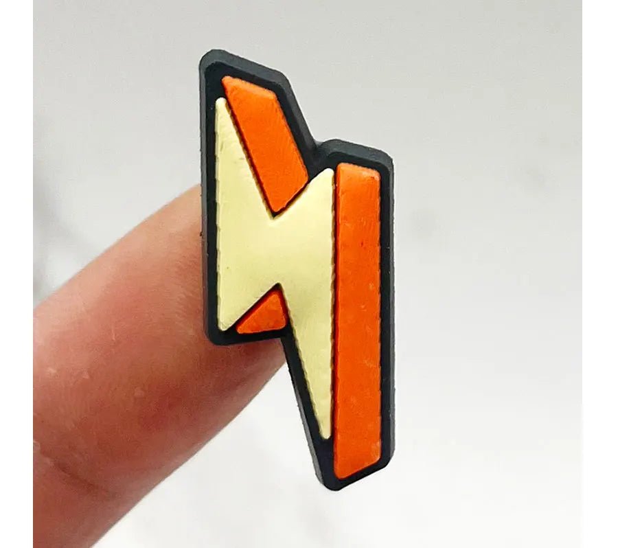 A Retro Roller Skate theme Shoe Charm, small and vintage, resembles a lightning bolt with black outlines and an orange-yellow design. It is held between two fingers against a plain background.
