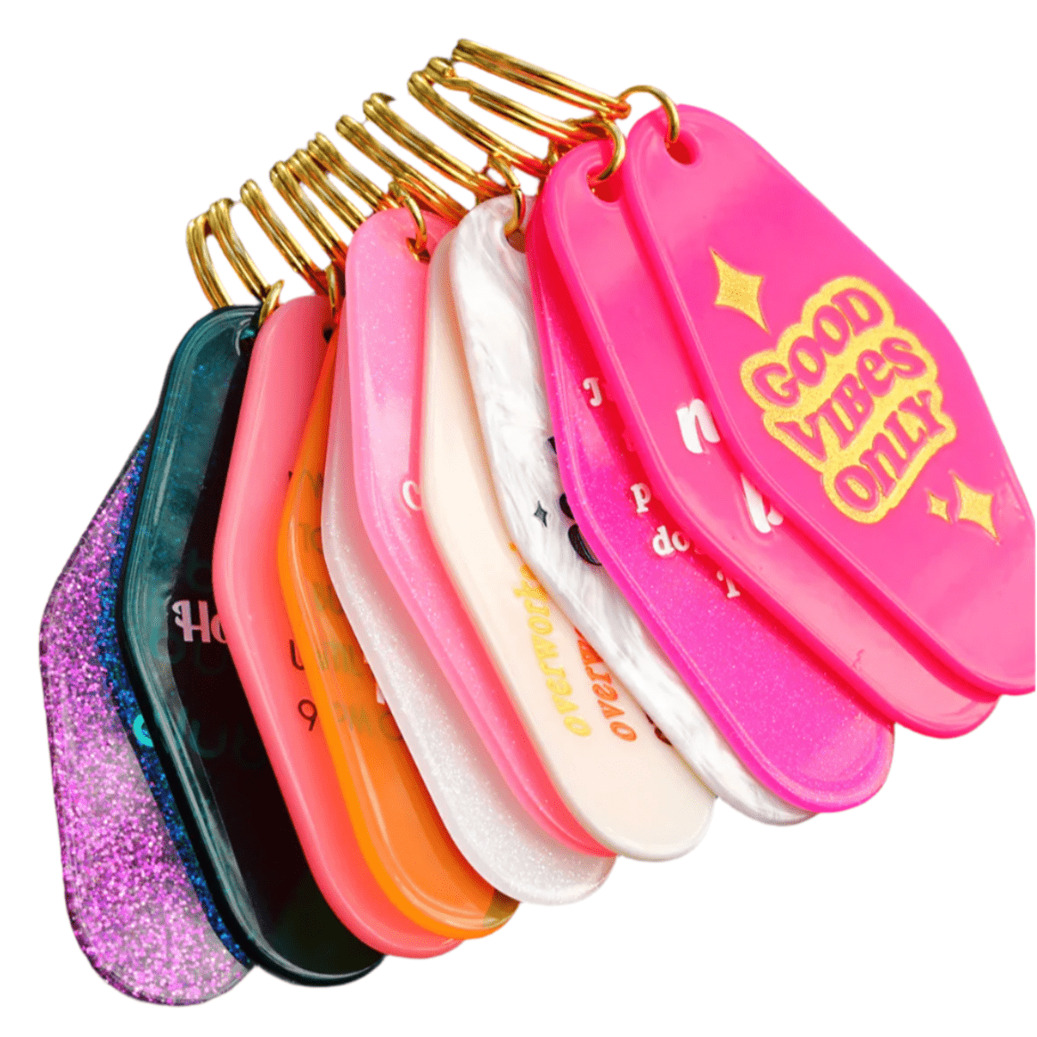A collection of Sassy Retro Motel Key Rings by Gay Skate hangs from golden loops. Each tag boasts shades of pink, orange, purple, and black—some with glitter. A 12cm key ring features "Good Vibes Only" in bright yellow lettering as a standout design choice.