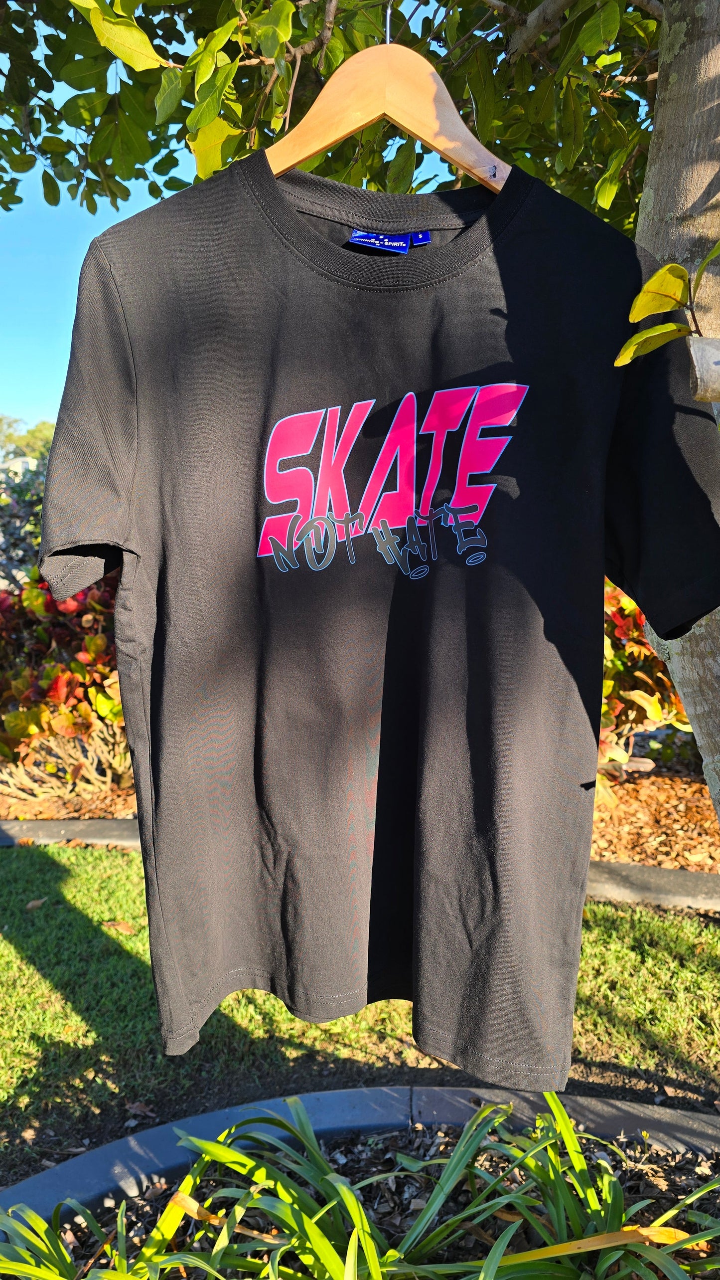 A My Store "Skate Not Hate - Local hand printed t-shirt" in black hangs on a wooden hanger outdoors in a garden setting. The shirt features the phrase "Skate Not Hate" in large, bold, pink and red gradient letters with a stylized, abstract design beneath it. Sunlight and shadows create a play of light on the fabric. Custom orders available.