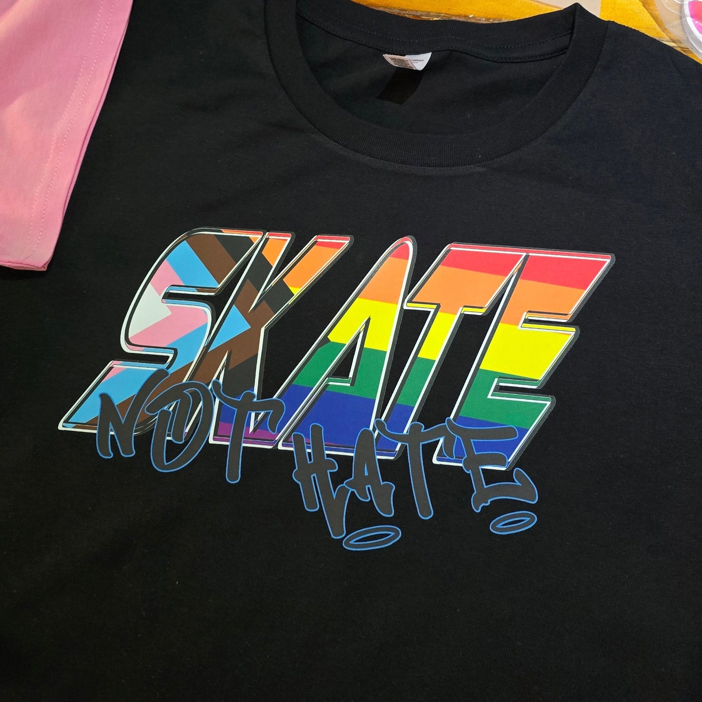 The SKATE NOT HATE original logo crop tee by Gay Skate is a white, short-sleeved shirt featuring the word "SKATE" in bold, pink, slanted capital letters across the chest. The message is accentuated with the word "NOT" in black, distorted text overlaying "SKATE," promoting the message: skate not hate.
