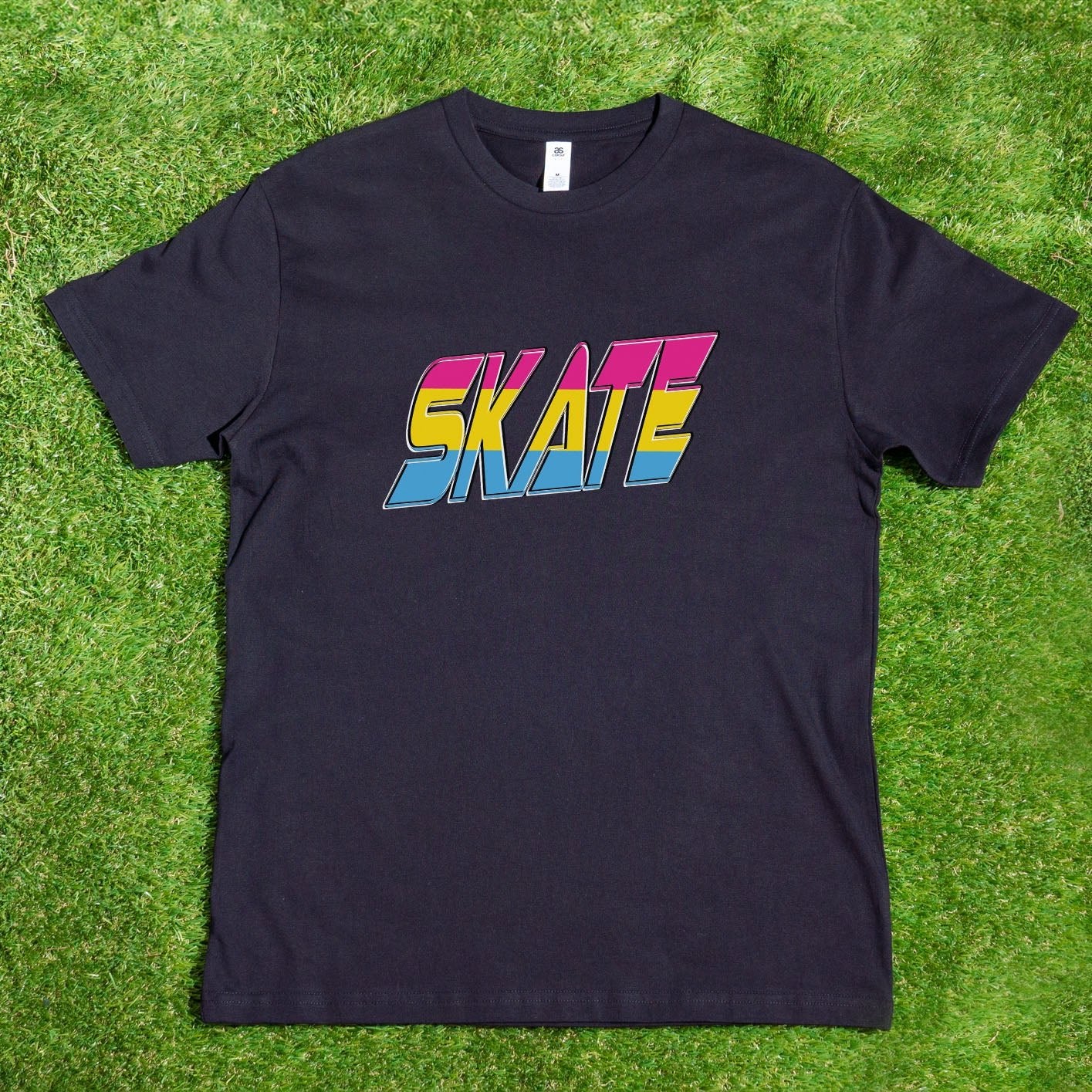 A SKATE Pansexual - Local hand printed t-shirt by My Store, featuring the word "SKATE" in bold, colorful letters with a 1980s retro style, is displayed on a wooden hanger from a tree branch. The scene includes lush greenery and vibrant flowers under sunlight casting beautiful shadows. Custom orders are available to celebrate diversity such as the Pansexual Pride Flag.