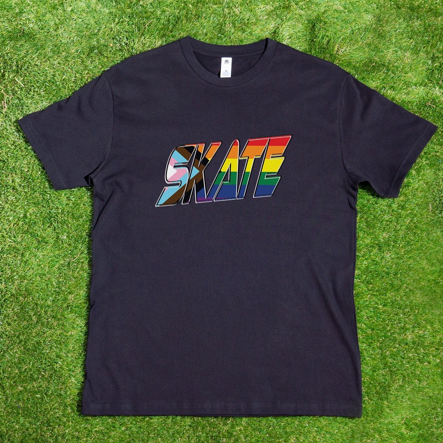 A SKATE Progress Rainbow Pride T-shirt from Gay Skate, featuring the word "SKATE" boldly printed in a vibrant rainbow gradient across the chest. This locally hand-printed AS Colour T-shirt is stylishly showcased on green grass, highlighting its striking design and color contrast.