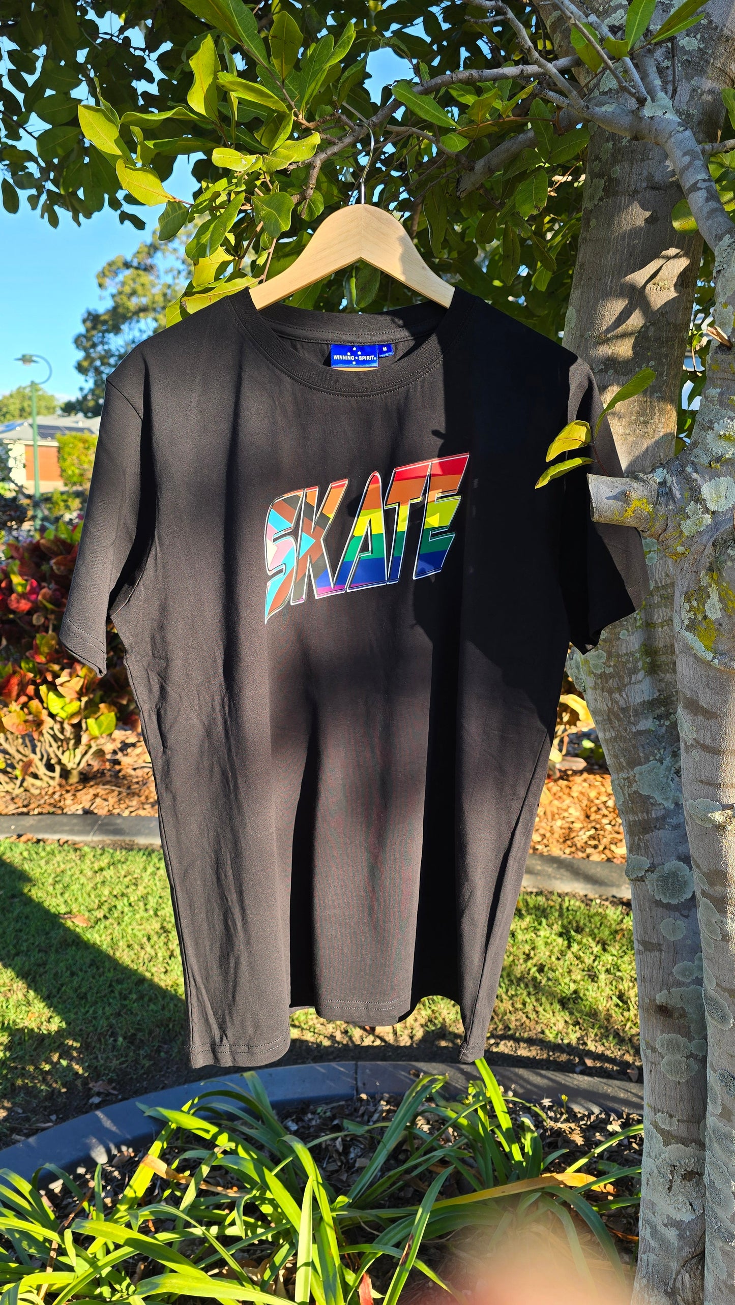A black SKATE Progress Pride - Local hand printed T-Shirt from Gay Skate hangs on a wooden hanger from a tree branch. The T-shirt showcases the word "SKATE" in large, colorful letters with a rainbow gradient and a glossy finish. The background features green foliage, a manicured garden, and a blue sky.