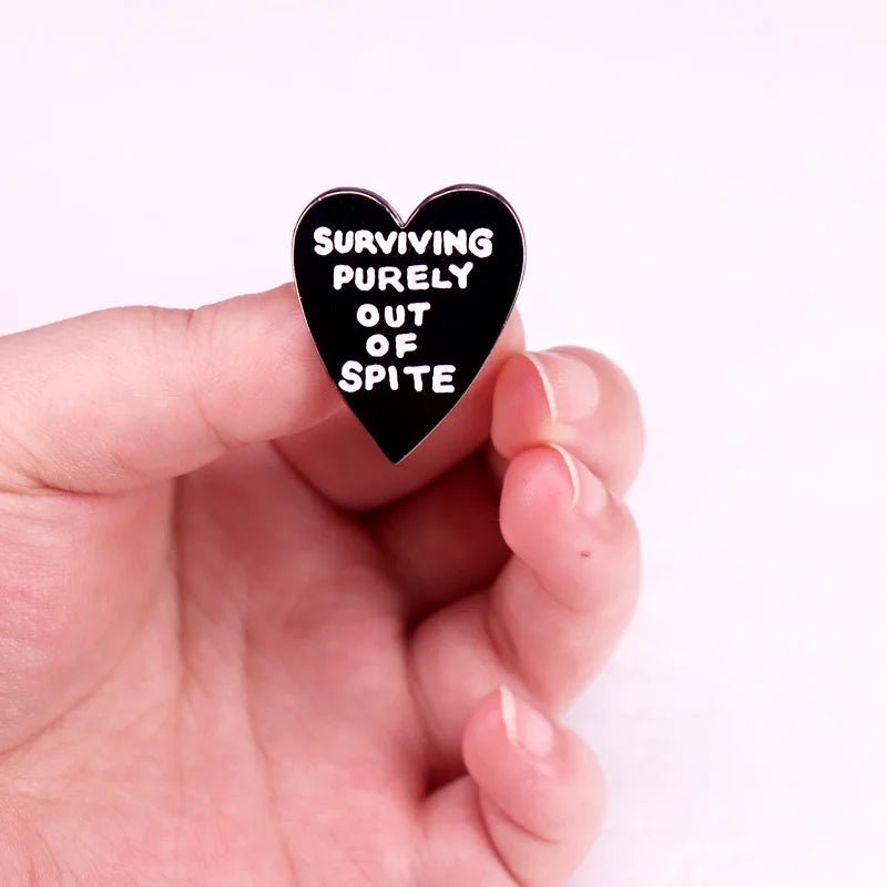 A hand holds the "Surviving Purely Out of Spite" black love heart badge with white text against a light pink background—an ideal bold pride accessory.