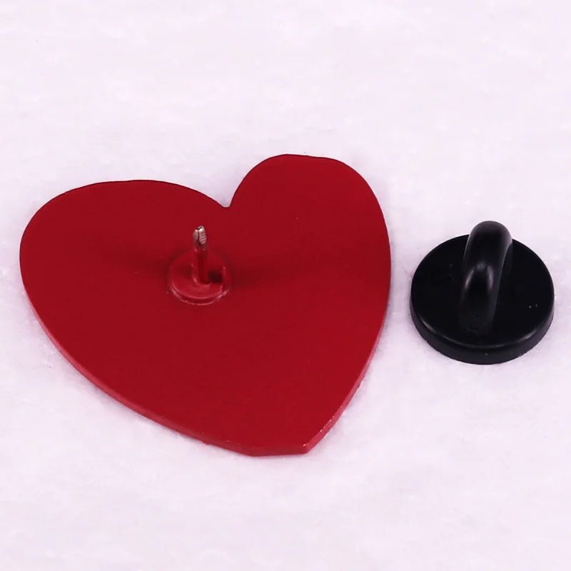 A "Too Cute To Be Neurotypical" pink heart enamel pin by Gay Skate and a black hook-shaped push pin lie on a white surface. The pink heart pin rests flat, showing the metal pin, while the black hook pin stands upright with its hook visible.