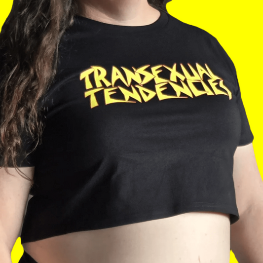 A person with long brown hair wears the "TRANSEXUAL TENDENCIES" local hand printed satire t-shirt, standing against a bright yellow background and expressing queer pride.