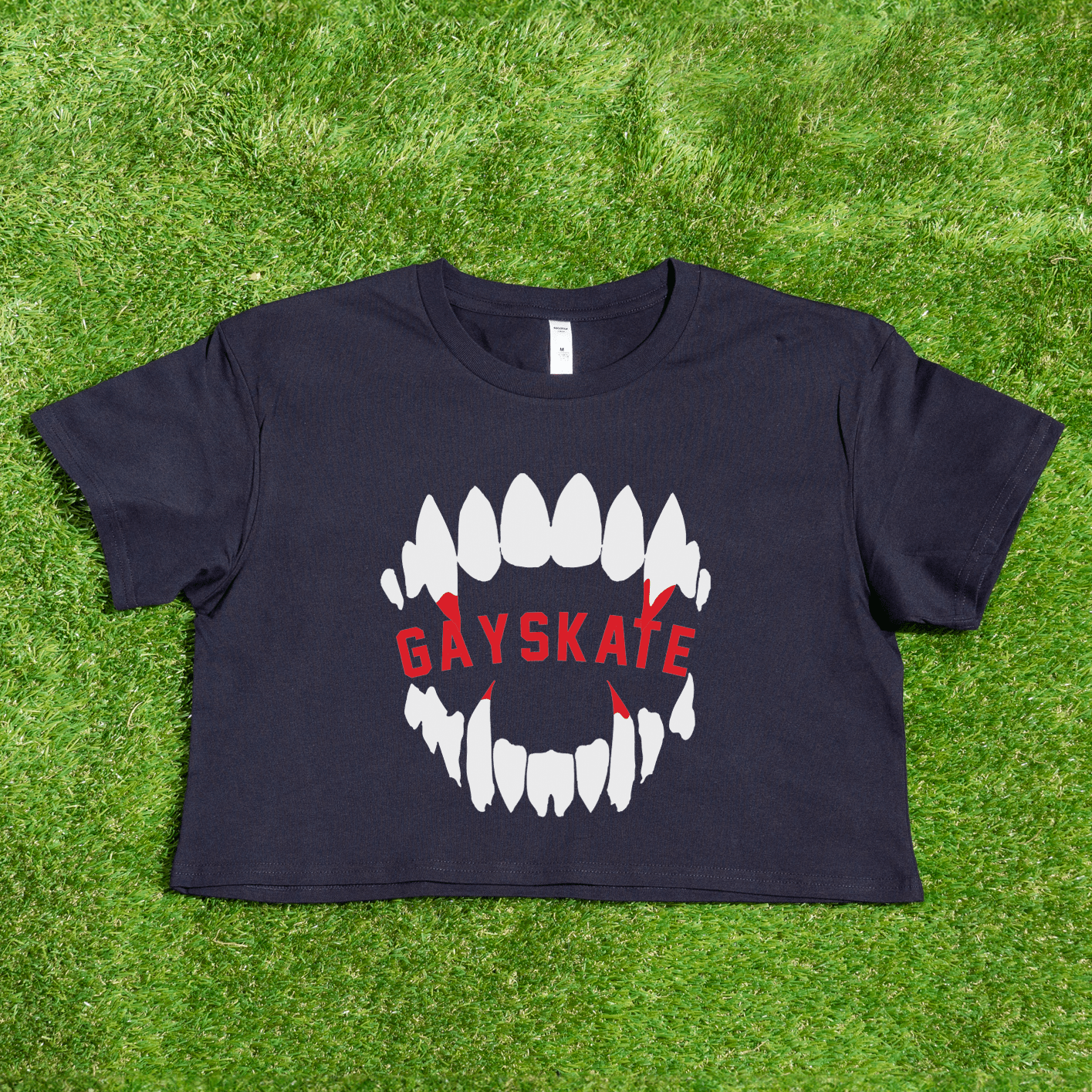 A black Vampire Skate - locally printed crop tee rests on grass, showing bold white teeth with red backdrops and "GAYSKATE" in red—perfect for fans of this queer-owned brand’s signature style.