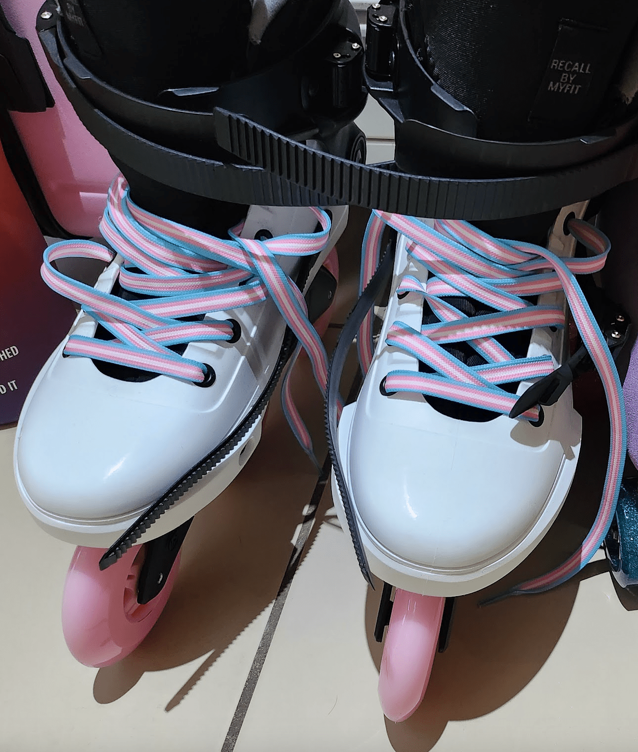 A pair of white rollerblades with pink wheels and Waxed Skate Laces in Non-Binary, Pansexual, Bisexual & Lesbian pride flag colors rest on a tiled floor.