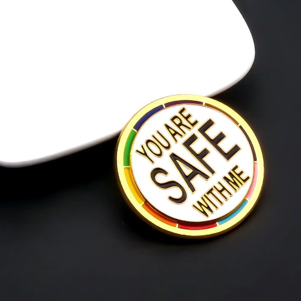 The "You Are Safe With Me - progress pride and gold round pin" features a gold border, rainbow accents, and bold black lettering—an empowering pride accessory displayed on a black surface beside a white object.