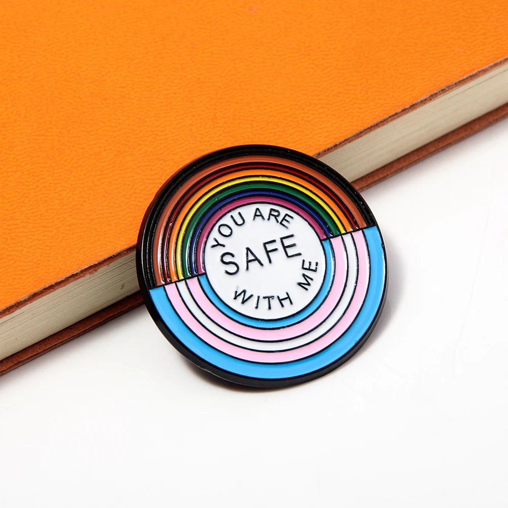 The "You Are Safe With Me" badge features rainbow and pastel stripes with the message in the center, ideal for pairing with pride key rings or other rainbow and trans pride ally badges, shown here on an orange notebook.