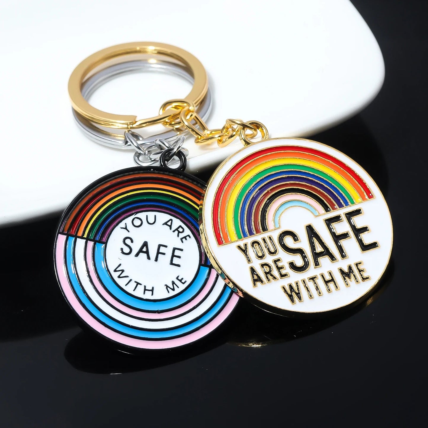 Two "You Are Safe With Me" key rings from the You Are Safe With Me rainbow and trans pride ally badges and key rings collection, featuring rainbow designs with gold rings. Ideal to pair with matching pride badges on display.