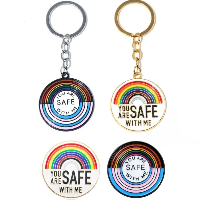Four round "You Are Safe With Me" pride badges and key rings with rainbow and trans designs: two as silver or gold key rings, two as charms only, each with unique fonts—perfect for showing allyship.