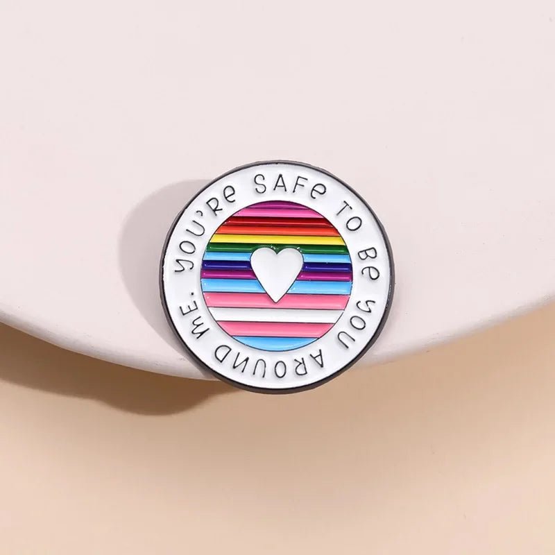 The "You're Safe To Be With Me" enamel ally badge is a round pin with a central heart encircled by horizontal rainbow stripes resembling the Rainbow Pride Flag. The text, "YOU'RE SAFE TO BE YOU AROUND ME," adorns the edge, ideal for pride accessory enthusiasts.