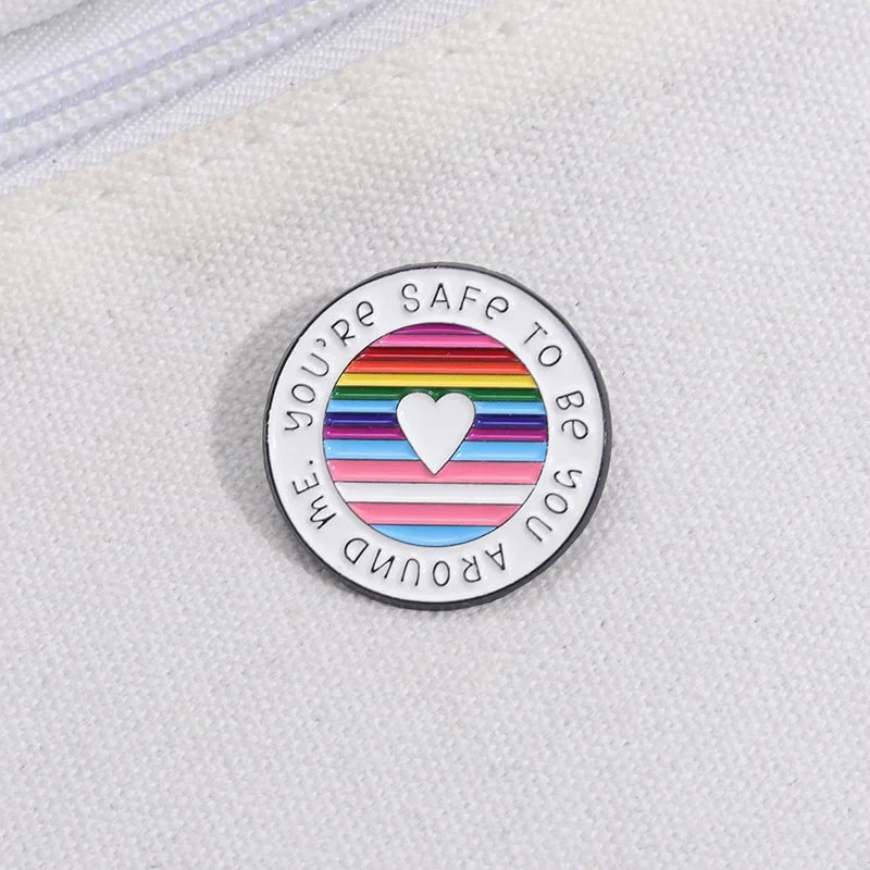 The You're Safe To Be With Me enamel ally badge is a round pin featuring a heart surrounded by rainbow stripes, reminiscent of a Rainbow pride flag. The text reads "YOU'RE SAFE TO BE YOU AROUND ME," and it rests on a white fabric background.