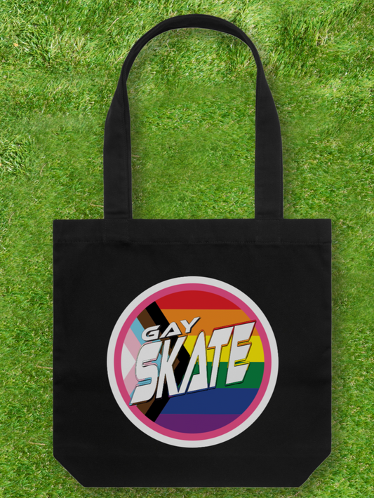 A large printed Cotton Canvas Tote Bag in black, featuring a striking GAY SKATE design with a circular logo on the front. The colorful emblem, set against rainbow stripes and intersecting black lines, represents a queer and trans-owned business. It rests beautifully on a green grass surface.