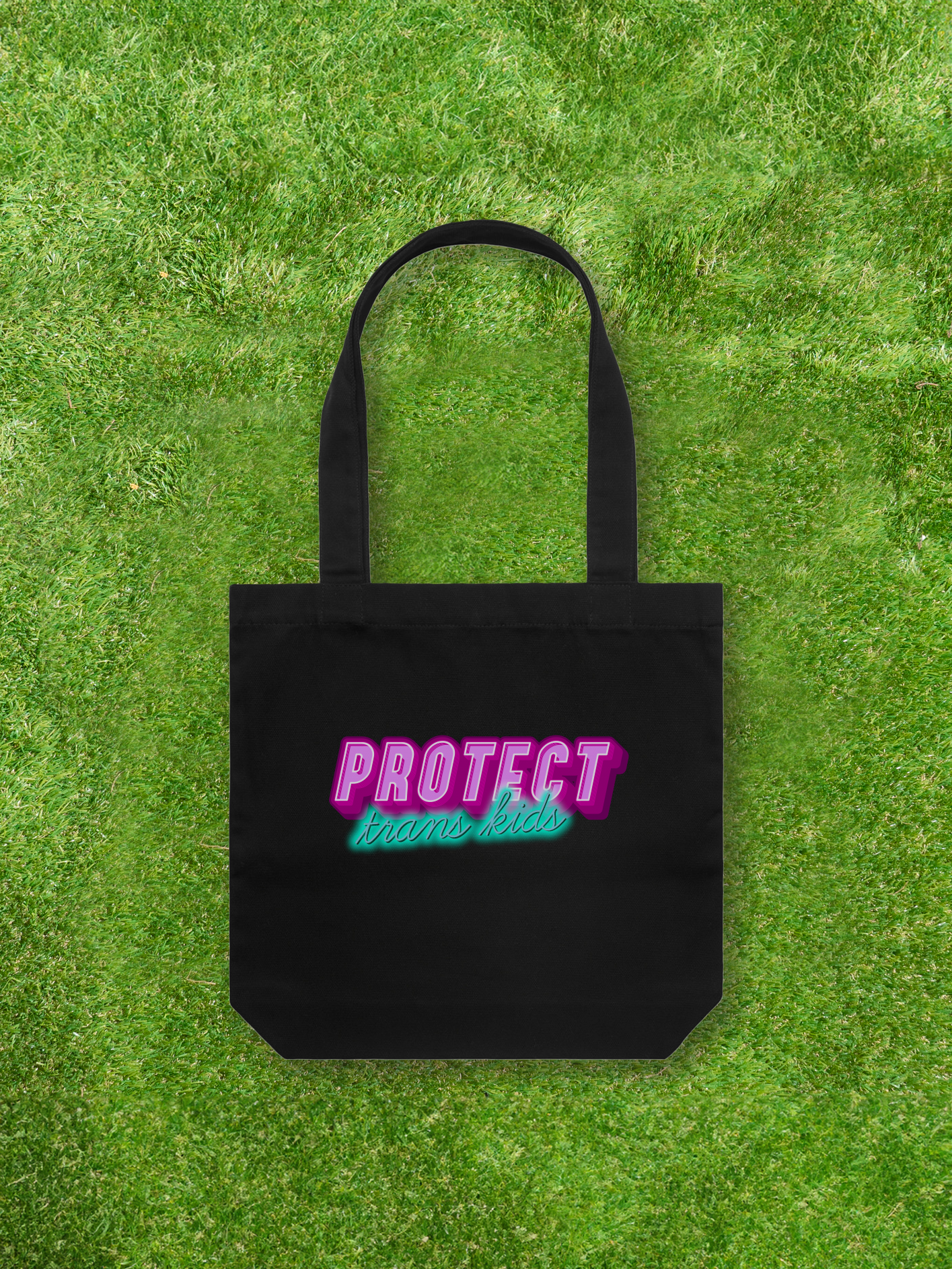 A Large printed Cotton Canvas Tote Bag - Black from Gay Skate, featuring pink and teal lettering that reads "Protect Trans Kids," rests on vibrant green grass, proudly representing a queer and trans-owned business.