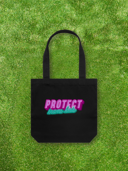 A Large printed Cotton Canvas Tote Bag - Black from Gay Skate, featuring pink and teal lettering that reads "Protect Trans Kids," rests on vibrant green grass, proudly representing a queer and trans-owned business.