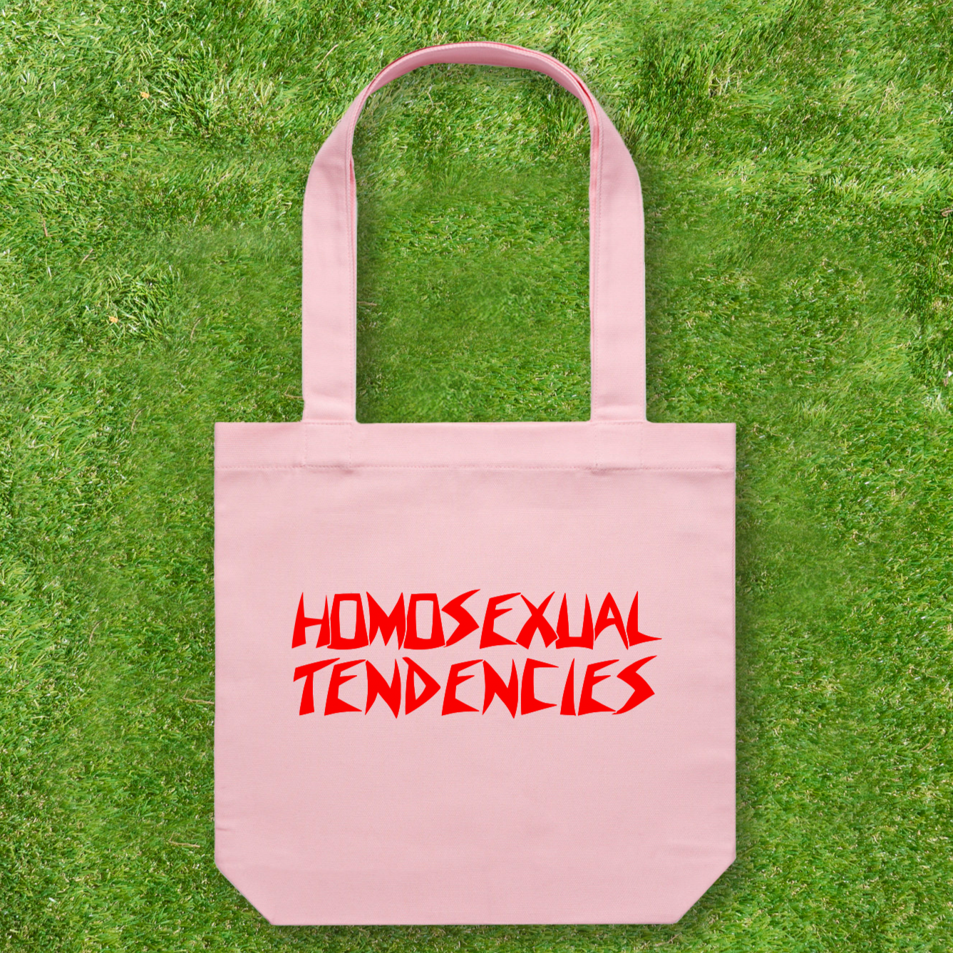 A Large printed Cotton Canvas Tote Bag in pink, featuring "Homosexual Tendencies" in bold red, angular letters, is designed by Gay Skate, a queer and trans-owned small business. This standout piece is set against a vibrant grass background.