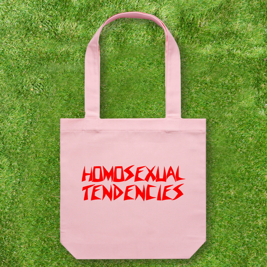 A Large printed Cotton Canvas Tote Bag from Gay Skate, proudly featuring "Homosexual Tendencies" in bold red letters, rests on green grass. This vibrant pink accessory is not just a statement piece but also supports a queer and trans-owned business.