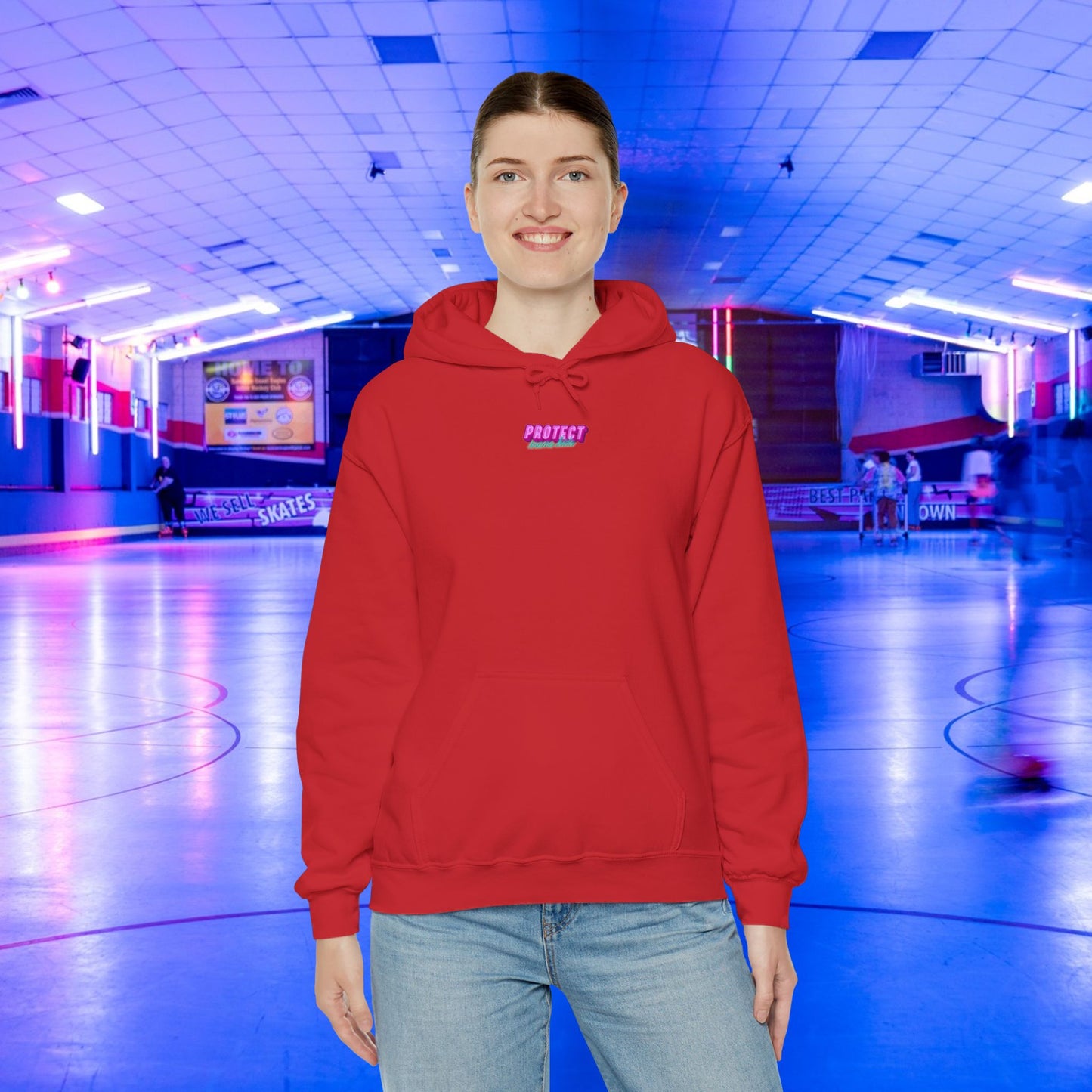 Wearing a "Protect Trans Kids" hoodie made from ethically grown cotton and jeans, a person smiles in an indoor roller skating rink with neon lights and a blue floor while motion-blurred skaters pass by.