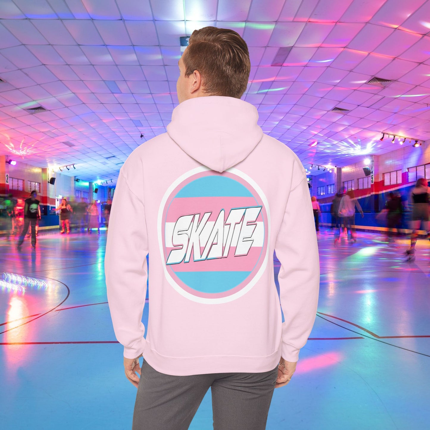 A person wearing a Printify SKATE Trans Flag round logo Hoodie, featuring the word "SKATE" inside a blue and pink circular graphic, stands at the center of a colorful roller skating rink with vibrant lights and other skaters in the background.