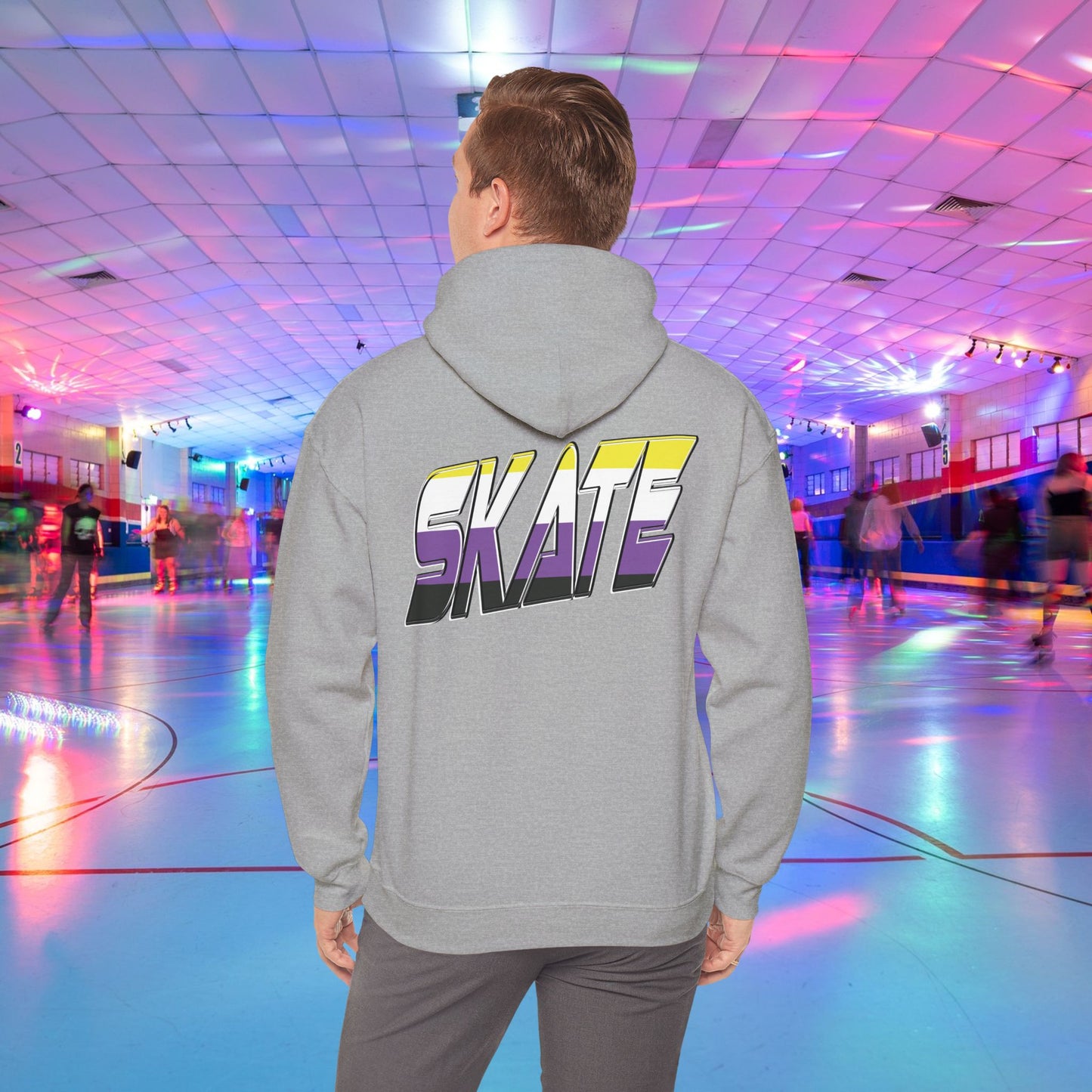 A person with light brown hair is wearing a high-quality SKATE Non-binary Pride Hoodie from Printify, featuring bold, colorful letters spelling "SKATE" on the back in non binary pride flag colours. They are standing in a roller skating rink with flashing neon lights, while other people skate around in the background.