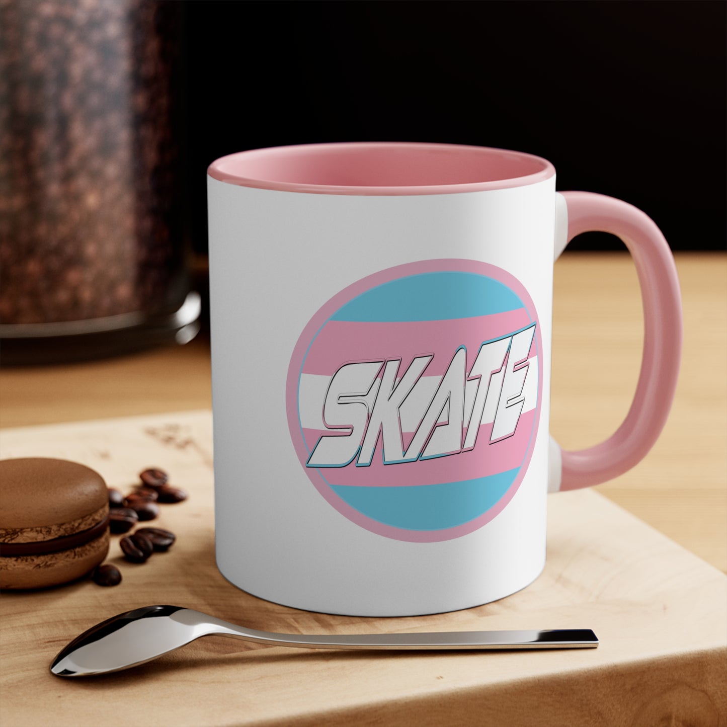 The SKATE trans flag circle design accent mug by Printify features a white exterior, blue handle, and blue interior. It showcases a circular design inspired by the trans flag, incorporating pink, light blue, and white colors. The word "SKATE" is prominently displayed in dynamic white letters at the center of the design.