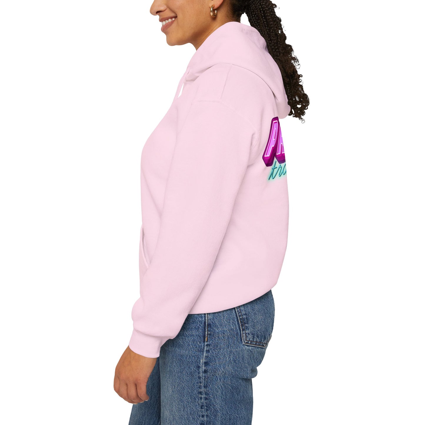 A person wearing a pink "Protect Trans Kids" unisex hoodie with vibrant back writing stands smiling, slightly turned to the side. Their curly hair is tied back, and they're in blue jeans against a plain white background.