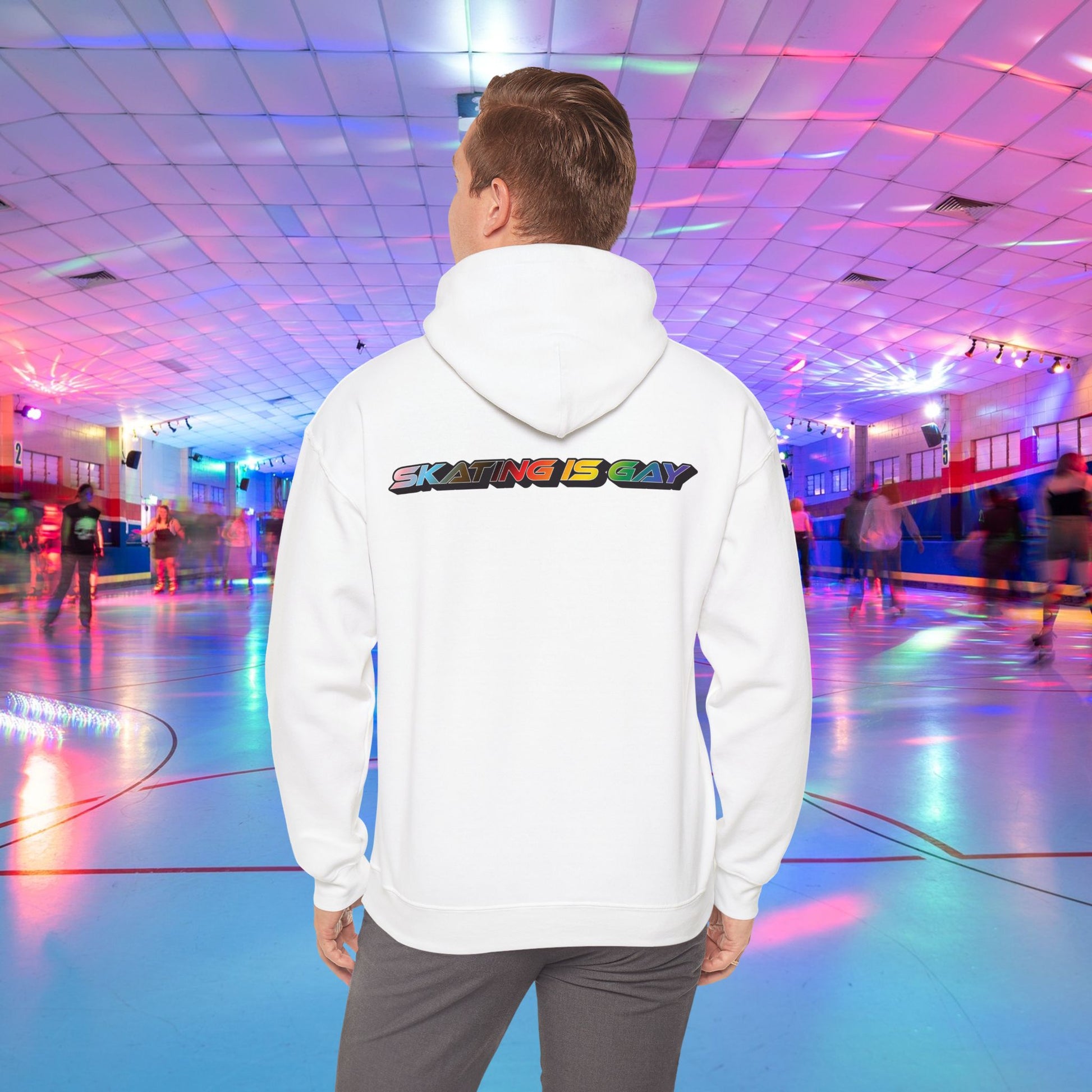 A person wearing a high-quality Skating Is Gay Hoodie by Printify, featuring the vibrant rainbow pride text "SKATING IS GAY" printed on the back, stands in the foreground of a neon-lit roller skating rink. People skate in the blurred background as colorful lights reflect off the rink's surface. The product is available with Australian shipping.