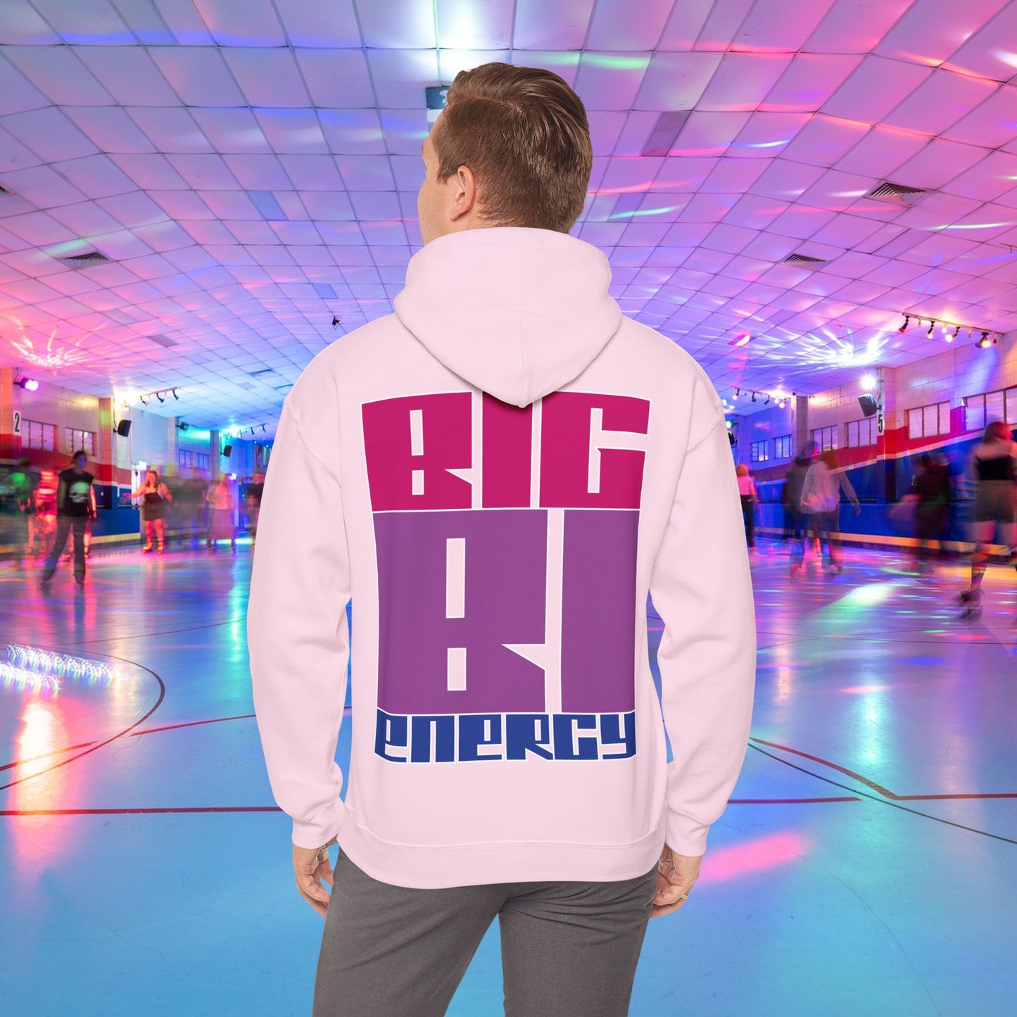 A person with light brown hair is wearing a high-quality BIG BI ENERGY Hoodie from GAY SKATE, featuring bold, Bisexual Pride Flag colour letters on the back. They are standing in a roller skating rink illuminated with vibrant, colorful lights. The cozy feel and warmth of this trans and queer-owned brand is evident.