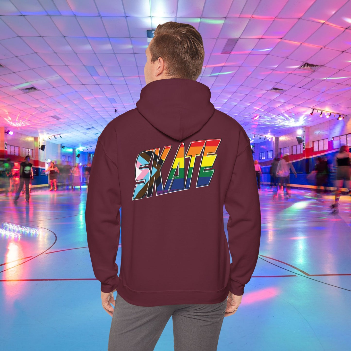 A person with short hair is wearing a GAYSKATE SKATE Progress Pride Hoodie, showcasing a rainbow pride flag gradient "SKATE" design on the back. They are facing away from the camera in a roller skating rink illuminated by vibrant, colorful lights and surrounded by other skaters.