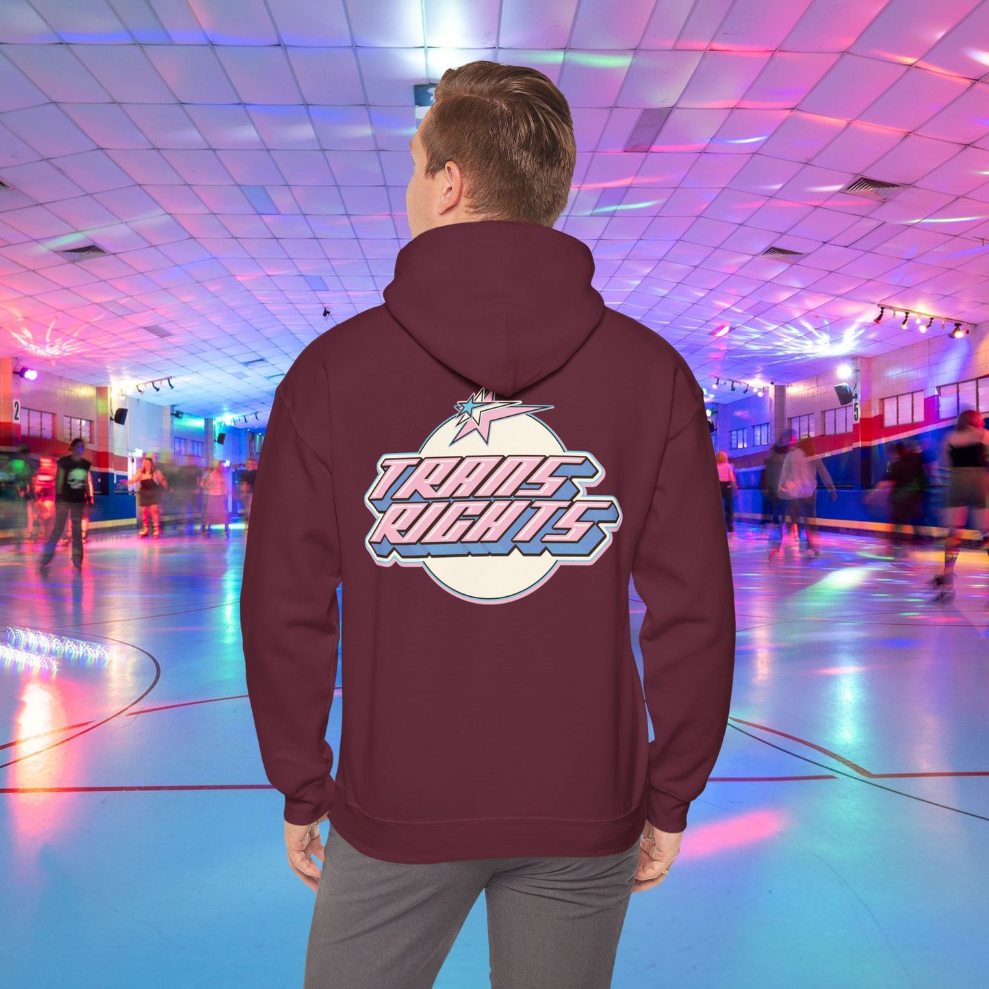 A person wearing a TRANS RIGHTS Hoodie in trans flag colours - Australian Shipping from GAY SKATE stands in a brightly lit roller skating rink, decorated with colorful lights and occupied by people skating in the background.