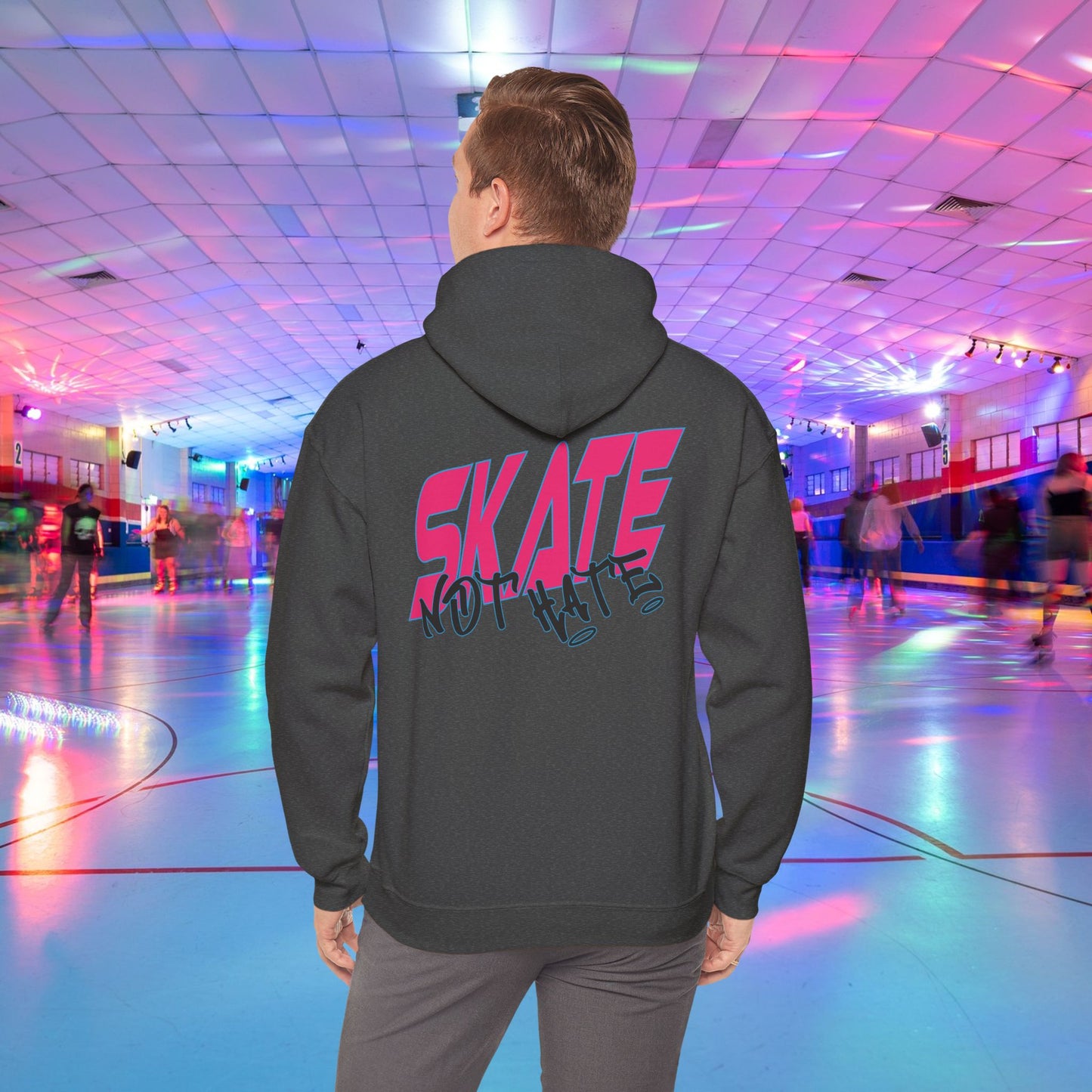 A person wearing a SKATE Not Hate Hoodie from Printify, characterized by its light pink color and high-quality construction, stands in a roller skating rink. The ethically-made unisex heavy blend hoodie, crafted from US-grown cotton, features the words "SKATE NOT HATE" printed on the back. The rink is adorned with colorful lights and has a shiny floor, with other skaters gliding in the background.