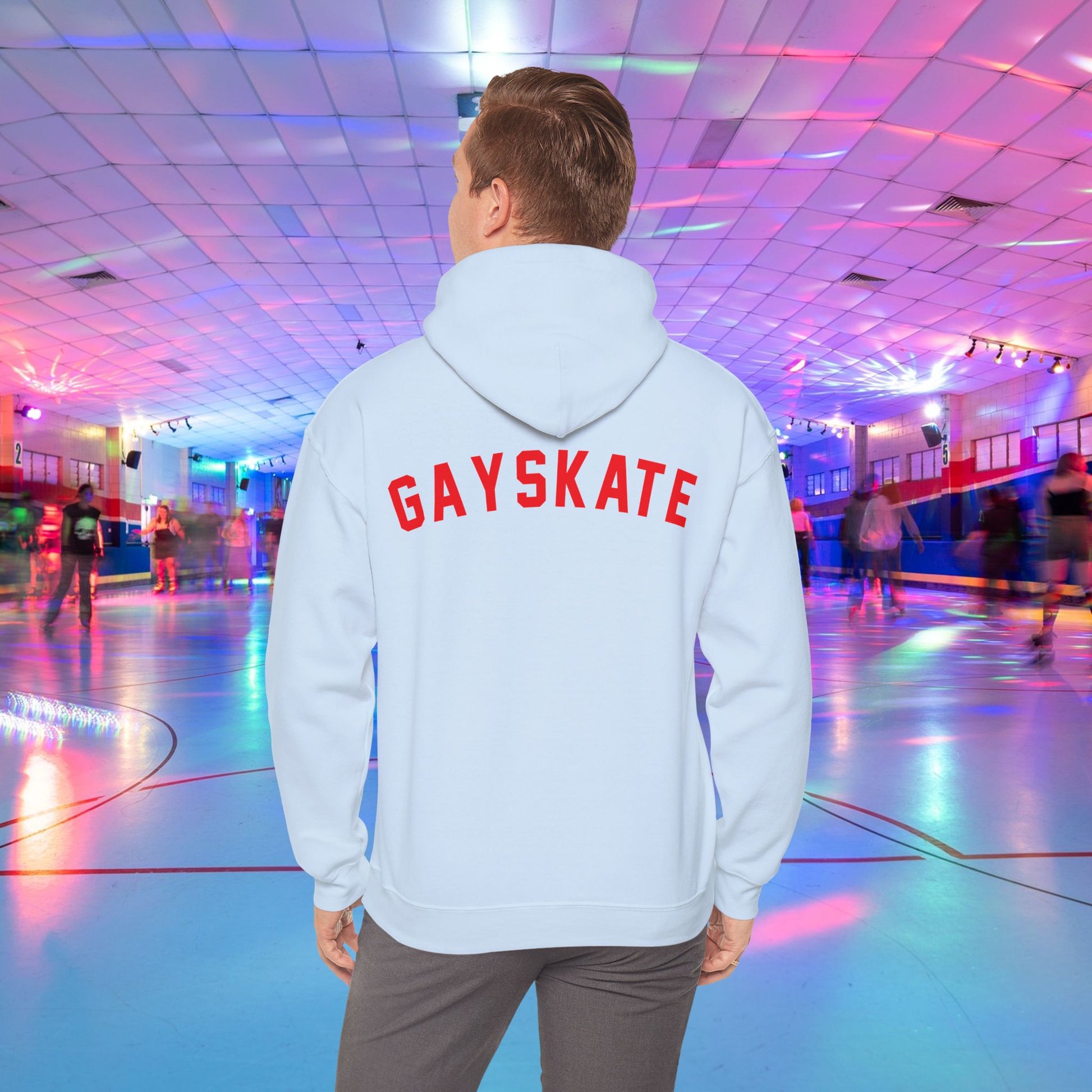 A person wearing a Printify Gay Skate 1982 Retro Hoodie, a high-quality unisex heavy blend hooded sweatshirt, with the word "GAYSKATE" in bold red letters on the back stands in a brightly lit roller skating rink. The rink features colorful lights and skaters in motion, creating a lively atmosphere.