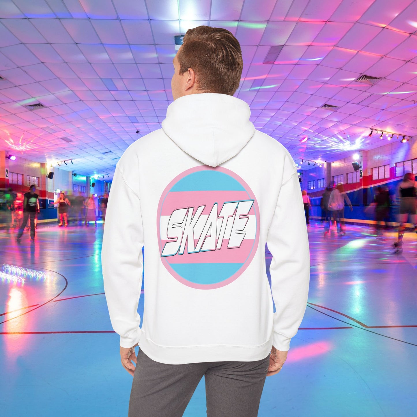 A person wearing a high-quality, unisex "Gay Skate Original - Trans Edition Hoodie" from Printify, featuring a large "SKATE" logo on the back, stands in a colorful roller skating rink. The rink is illuminated with multicolored lights and has several people skating in the background. The overall atmosphere is lively and vibrant.