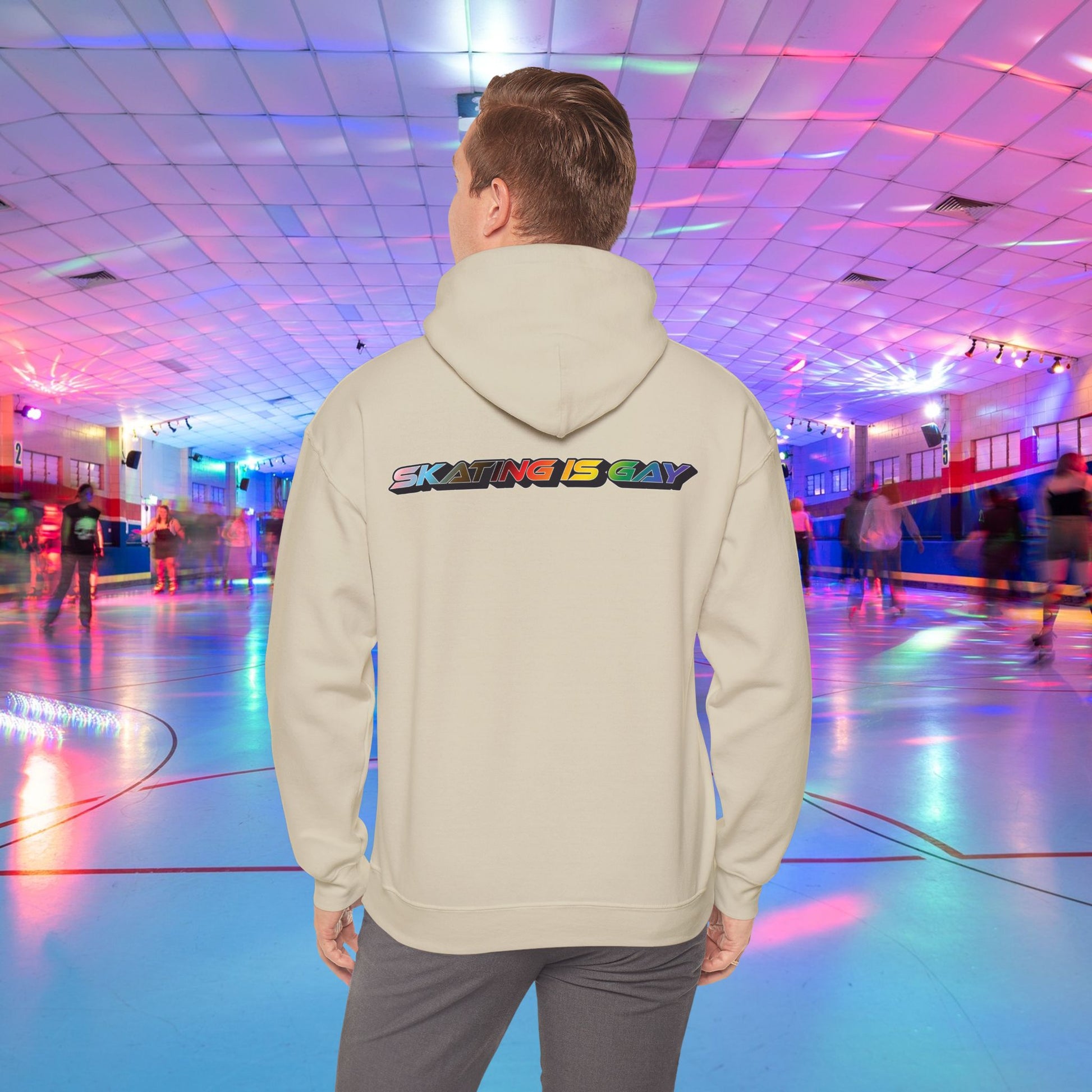 A person wearing a high-quality Skating Is Gay Hoodie by Printify, featuring the vibrant rainbow pride text "SKATING IS GAY" printed on the back, stands in the foreground of a neon-lit roller skating rink. People skate in the blurred background as colorful lights reflect off the rink's surface. The product is available with Australian shipping.