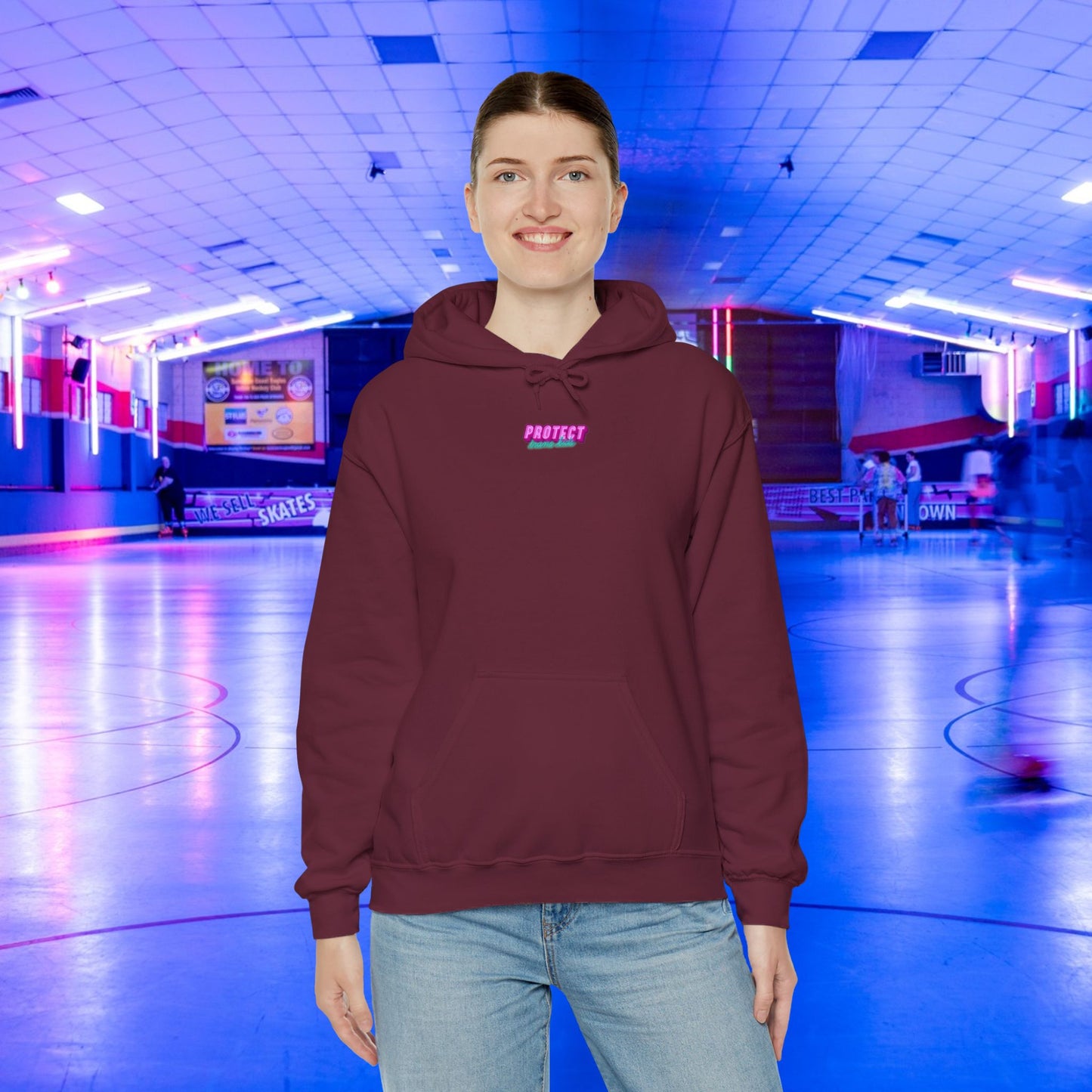 A smiling person stands in a roller skating rink, wearing the Protect Trans Kids Hoodie—a maroon, unisex hoodie made from ethically grown cotton—and jeans. The background glows with blue and pink neon lights as skaters glide by.