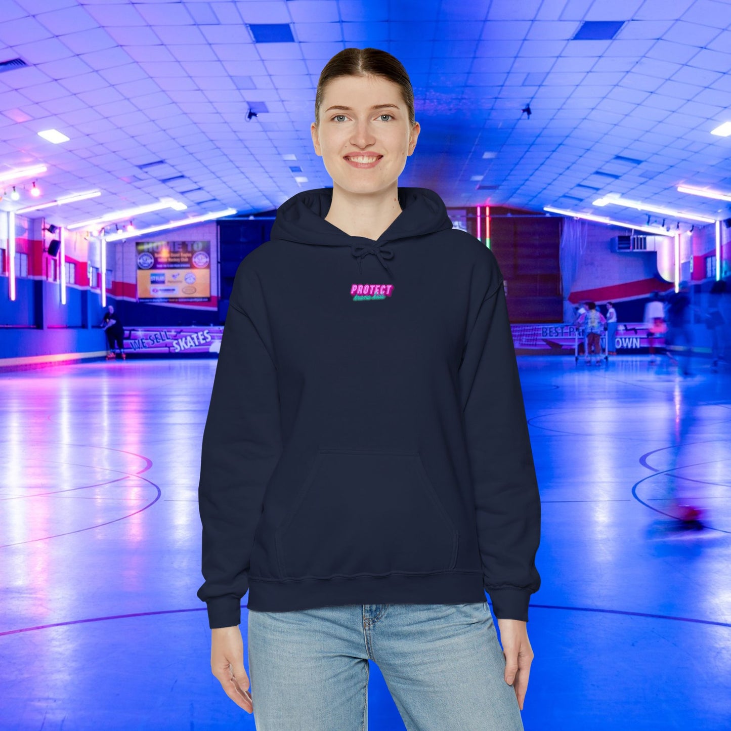 In a roller skating rink, a person dons the "Protect Trans Kids Hoodie" in navy, made from ethically grown cotton. The colorful and vibrant rink is filled with skaters, creating an energetic atmosphere.